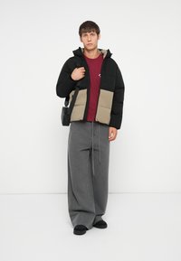 Padded jacket with black and beige sections, hood, and zip closure. Maroon t-shirt beneath. Grey wide-leg pants and black footwear.