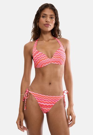 Femme portant un bikini halter à motif zigzag rose et rouge avec un bas à nouer sur les côtés, debout devant un fond clair uni.