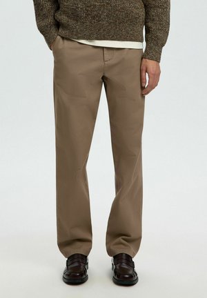 Man met een bruine broek met rechte pijpen, donkerbruine leren schoenen en een bruine gebreide trui met handen langs het lichaam.