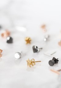 Verschiedene Steckerohrringe mit Silber-, Gold- und Schwarz-Tönen in verschiedenen Formen, darunter Kreise, Herzen und Sterne, auf einem hellen Hintergrund.