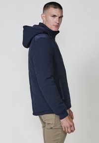 Felpa con zip di colore blu scuro, realizzata in un tessuto strutturato, con un ampio cappuccio e tasche laterali. Design aderente con polsini e bordo a costine.