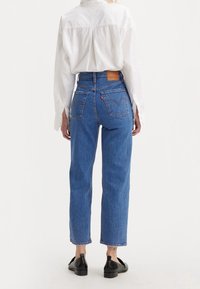 Jeans de mezclilla azul de talle alto con corte recto, con bolsillos traseros, un parche de cuero y costuras en contraste. Llevados con una camisa blanca.