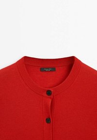 Cardigan rouge en maille avec encolure ronde et boutons noirs, présenté des épaules au haut de la poitrine sur fond blanc uni.