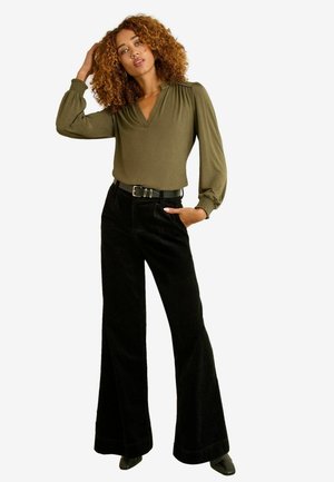Olivgrüne Bluse mit Puffärmeln und V-Ausschnitt, kombiniert mit schwarzen Cord- Schlaghosen und einem schwarzen Gürtel. Glatte Textur und figurbetontes Design.