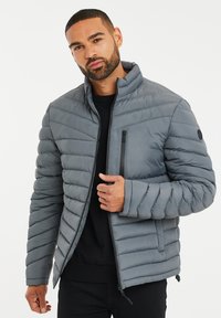 Grå puffjakke med høj krave, diagonalt quiltet mønster, frontlynlås og en lynlåslomme på brystet. Tekstureret, letvægtsmateriale.