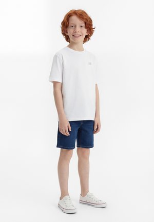 Červenovlasý chlapec v bielom tričku s krátkym rukávom, modrých džínových šortkách a bielych teniskách stojí a usmieva sa na bielom pozadí.