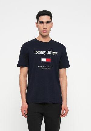 Bărbat în picioare, îmbrăcat cu un tricou Tommy Hilfiger de culoare bleu marin, cu logo-ul și textul "Established MCMLXXXV New York City" pe față.