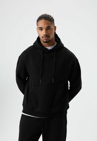 Zwarte hoodie gemaakt van zacht materiaal, voorzien van een kangoeroezak, trekkoorden en een losse pasvorm. Draagt met bijpassende zwarte broek.