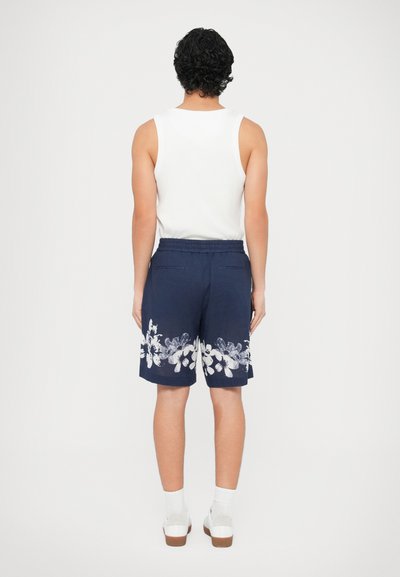 Shorts bleu marine avec motif floral blanc, taille élastique et poches latérales; portés avec un haut sans manches blanc et des chaussures blanches.