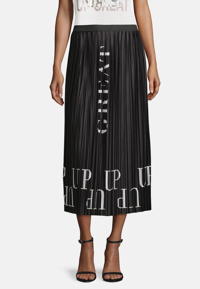 Jupe midi plissée noire avec texte blanc "UP" imprimé verticalement et horizontalement, portée avec des talons noirs à bride chevillante, bas du torse visible.
