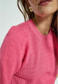Rosa gestrickter Pullover mit einem strukturierten Muster und geripptem Kragen, kombiniert mit goldenen blumigen Ohrringen. Nahaufnahme, die auf das Design fokussiert ist.