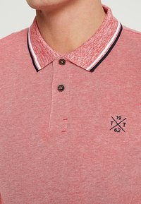 Červené polo tričko s texturovanou tkaninou, kontrastním límcem s námořnickými a bílými pruhy, dvěma knoflíky a malým emblémem s překříženými liniemi.