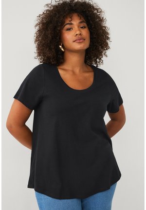 Kvinde med krøllet hår iført en sort kortærmet top med rund halsudskæring og blå jeans, stående med hænderne bag ryggen.