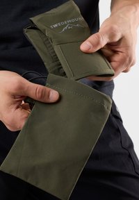 Pochette di tessuto verde con una superficie liscia e bordi cuciti, caratterizzata da un logo nella parte superiore. Tenuta in mano vicino a un abbigliamento scuro.