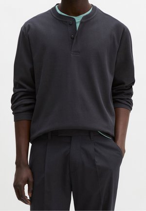 Homme portant une chemise henley manches longues gris foncé sur un sous-vêtement bleu clair avec un pantalon foncé, une main dans la poche.