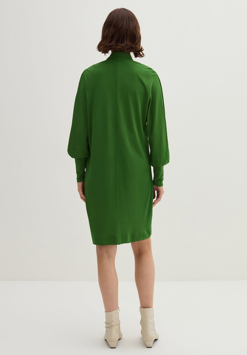 Groene satijnen jurk met lange mouwen en een hoge kraag. De achterkant laat een gladde textuur en een iets losse pasvorm zien, die stopt boven de knie.