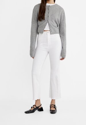 Femme portant un cardigan gris court, un jean blanc taille haute et des chaussures Mary Jane noires à petit talon, debout sur fond blanc.