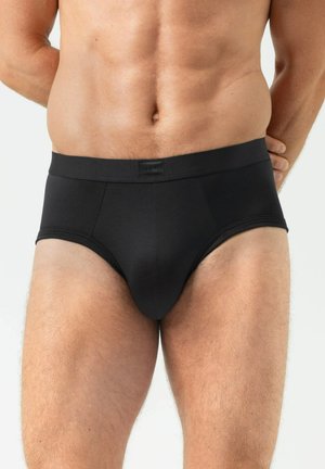 Männlicher Oberkörper in schwarzer Unterhose, zeigt definierten Bauch und Oberschenkel vor hellem einfarbigem Hintergrund.