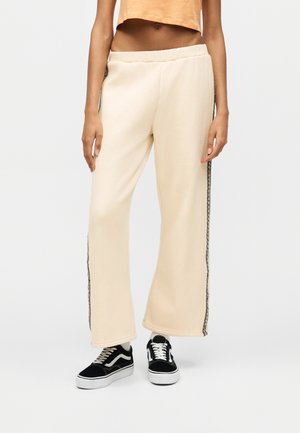 Άτομο που φοράει κρεμ παντελόνια με φαρδιά πέτα και πλαϊνές ρίγες και μαύρα και άσπρα αθλητικά παπούτσια, στέκεται μπροστά από απλό φόντο.