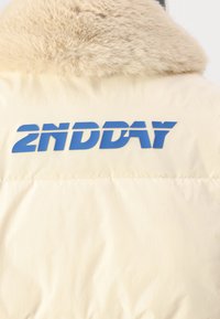 Puffer jacket in cream with a faux fur collar. Features a prominent blue graphic text "2NDDAY" across the back. Smooth texture, quilted design.