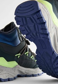 Buty trekkingowe w ciemnym kolorze z zielonymi akcentami, solidna gumowa podeszwa oraz teksturowana szara środkowa podeszwa. Wyraźny wzór bieżnika zapewniający przyczepność.