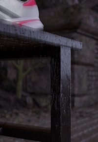 White and pink sneaker resting on the edge of a dark wooden bench with a blurred stone and tree background.