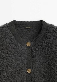 Cardigan en maille texturée gris foncé avec encolure ronde et deux boutons dorés brillants sur le devant, étiquette visible à l'intérieur du col.