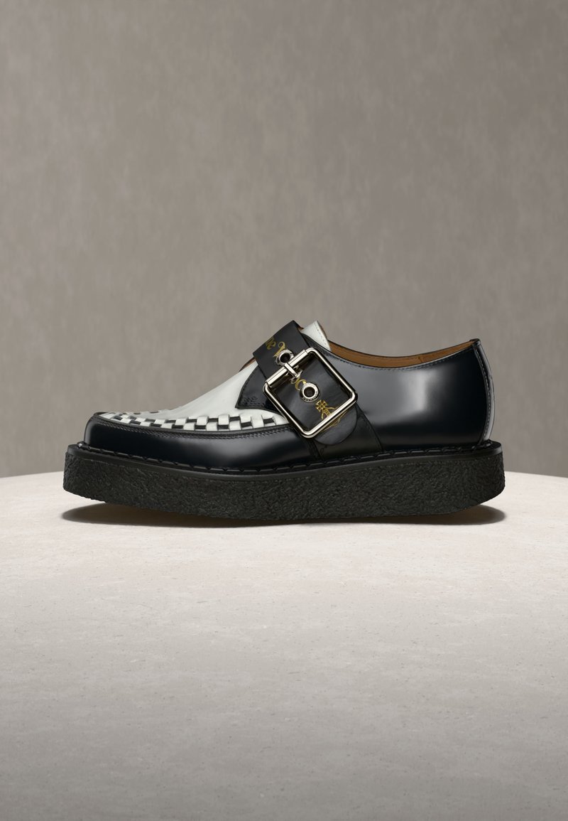 Schwarz-weißer Lederschuh mit geflochtenem Zehendesign, großem silbernem Schnallenriemen und dicker strukturierter schwarzer Sohle auf neutralem Untergrund.