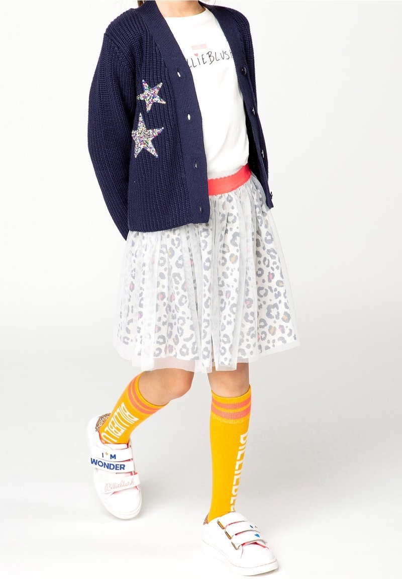 Marineblauwe cardigan met twee accenten van pailletten sterren, wit T-shirt, doorschijnende witte rok met luipaardprint en kniehoge gele sokken met strepen. Witte sneakers.