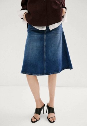 Kvinde iført en knælængde denimnederdel, mørkebrun top over en hvid skjorte og sorte højhælede sandaler med stropper.