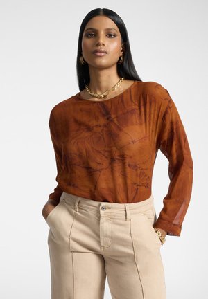 Frau mit langen schwarzen Haaren, die eine braune Batikbluse und beige Hose trägt, ergänzt durch goldene Creolen, eine Halskette und Armbänder.