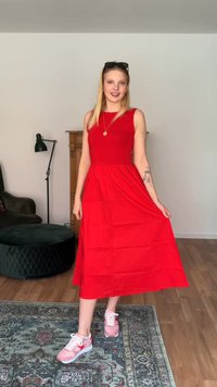 Femme debout à l'intérieur portant une robe rouge sans manches, des baskets roses, des lunettes de soleil sur la tête et un collier pendentif, tenant l'ourlet de sa robe.