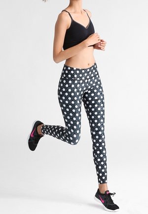 Soutien-gorge de sport noir associé à des leggings moulants avec un motif à pois blancs sur fond noir. Le modèle porte des baskets noires.