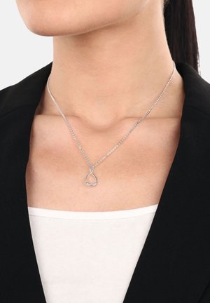 Collana in argento a forma di infinito con piccole pietre incastonate, indossata attorno al collo sopra una maglietta bianca e un blazer nero.