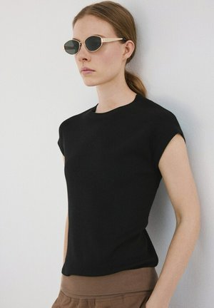 Ženska s svetlo rjavimi lasmi, spetimi v čop, ki nosi ovalna črna sončna očala, črno brezrokavnik in rjave hlače, stoji ob beli steni.