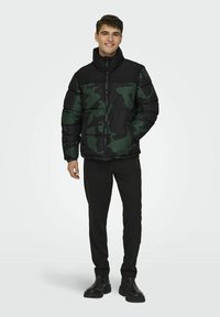 Giacca puffer verde e nera a motivo camouflage con colletto alto, zip frontale e polsini a costine, indossata con pantaloni neri e stivali.