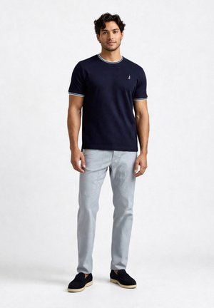Uomo in piedi con maglietta blu navy con bordi bianchi, pantaloni grigio chiaro e scarpe slip-on nere su sfondo bianco semplice.