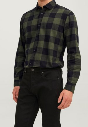 Homme portant une chemise à manches longues à carreaux verts et noirs, rentrée dans un pantalon noir, sur un fond clair uni.