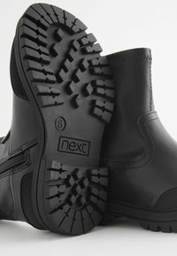 Bottines noires avec une semelle en caoutchouc texturé, détail de fermeture éclair sur le côté, tige en cuir lisse et coutures renforcées sur les bords.