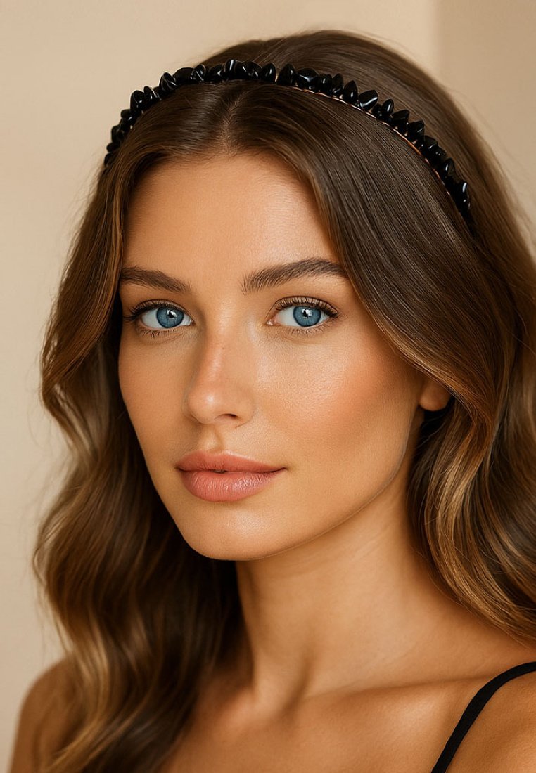 Czarna opaska na głowę z ostro zakończonymi detalami, o połyskującym wykończeniu. Gładkie, falujące włosy opadają na ramiona, podkreślając niebieskie oczy i neutralny kolor ust.