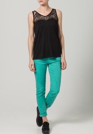 Top nero senza maniche con design in pizzo, abbinato a pantaloni aderenti turchesi e sneakers nere. Sfondo grigio neutro.