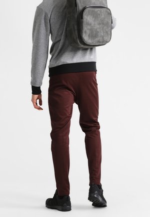 Persona che indossa pantaloni color amaranto, scarpe da ginnastica nere, una felpa grigia con polsini neri e uno zaino Adidas grigio, che cammina via su uno sfondo bianco semplice.