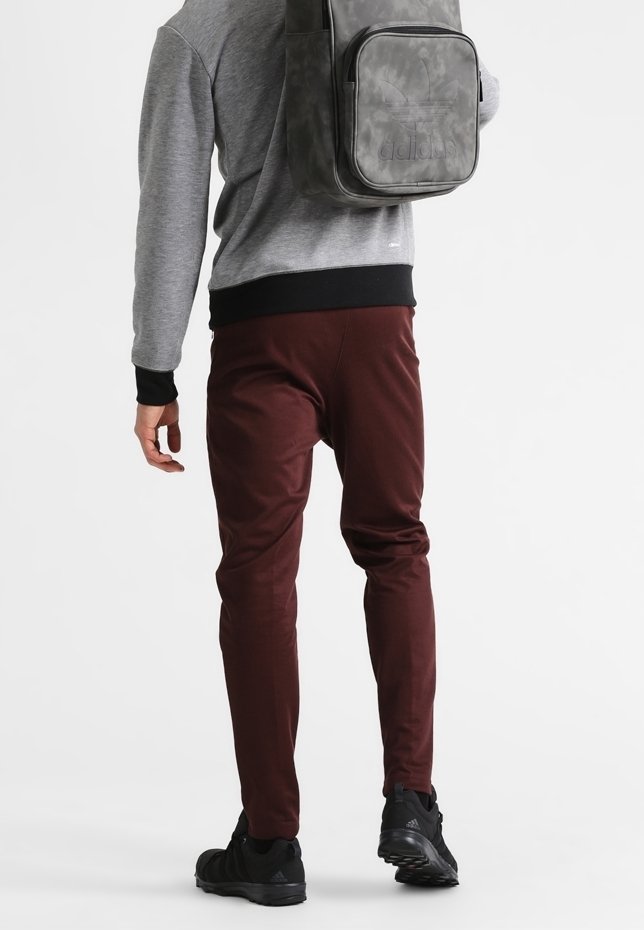 Osoba ubrana w bordowe spodnie, czarne trampki, szarą bluzę z czarnymi mankietami oraz szary plecak Adidas, idąca odwrócona na jednolitym białym tle.