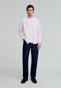 Cămașă roz deschis, cu butoni, purtată peste un tricou alb, asociată cu blugi închis la culoare, lejeri și pantofi negri. Detaliu simplu brodat prezent.