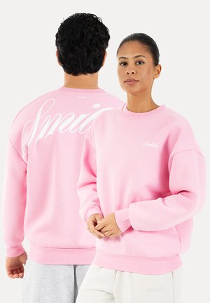 Deux personnes portent des sweat-shirts rose pastel assortis ; l'une fait face avec les mains jointes, l'autre montre un grand texte blanc dans le dos.
