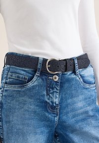 Jean bleu avec une ceinture noire à boucle argentée présentant un motif léopard subtil, portée avec une chemise blanche à manches longues, détails de la taille et des hanches visibles.