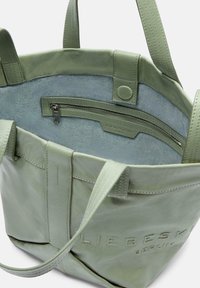 Zelená kožená tote taška s texturovaným povrchem, obsahující zipovou kapsu a kontrastní světle modrou látkovou podšívku. Ražené logo na přední straně.