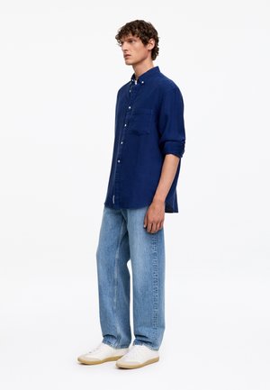 Homme debout portant une chemise bleu foncé à boutons avec les manches retroussées, un jean bleu clair et des baskets blanches sur fond blanc.