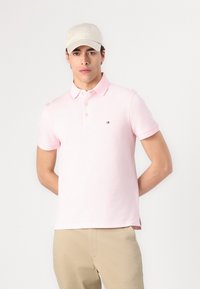 Camisa polo rosa de manga corta con tapeta de tres botones. Presenta un pequeño logo en el pecho. Se lleva con pantalones beige y una gorra color crema.