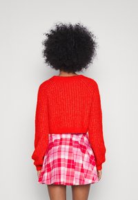 Roter gestrickter Pullover mit langen Ärmeln und einem cropped Design, kombiniert mit einem rosa karierten Faltenrock, der unterschiedliche Schattierungen von rosa Quadraten aufweist.